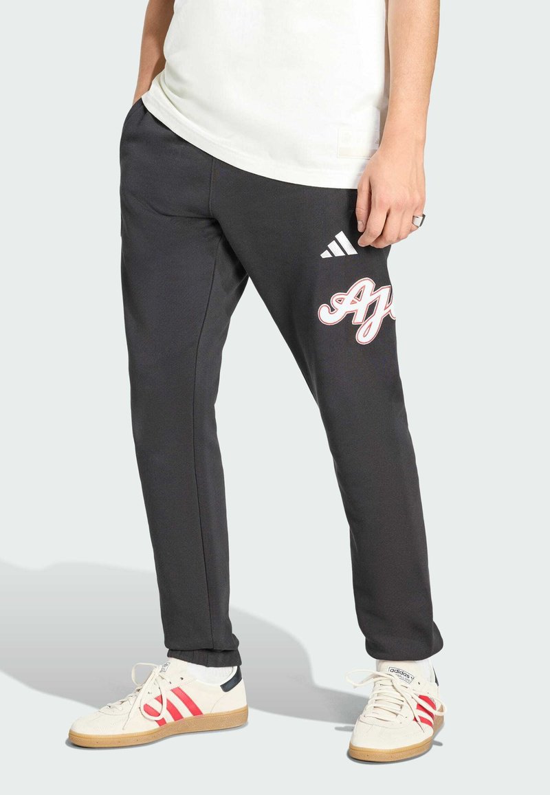 Persona che indossa pantaloni della tuta neri con logo bianco e rosso sulla coscia, sneakers bianche con strisce rosse e maglietta bianca.