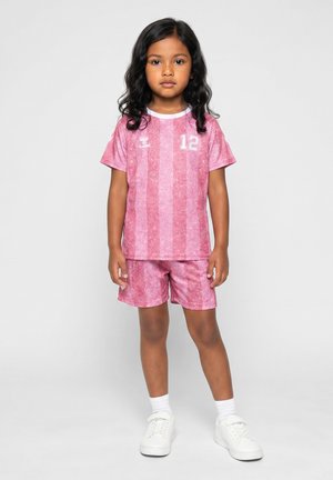 L’enfant se tient de face, vêtu d’un maillot de sport rose rayé avec le numéro 12 et d’un short assorti, avec des chaussettes blanches et des baskets blanches.