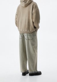Sudadera beige de corte relajado, combinada con jeans holgados de color verde claro, con costuras visibles y dos bolsillos traseros. Unos zapatos negros completan el look.