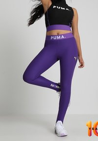 Fioletowe legginsy ze strukturą, z wysokim stanem i logo. Czarna bezrękawnik z białym brandingiem. Białe buty sportowe z fioletowymi akcentami.