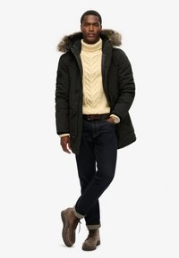 Zwarte gewatteerde jas met een bontgevoerd capuchon, crèmekleurige kabeltrui, donkere spijkerbroek en bruine veterschoenen. Casual winteroutfit.