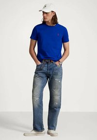 Jeune homme portant une casquette blanche, un t-shirt bleu roi, un jean bleu déchiré avec une ceinture rayée, des baskets blanches et une montre, se tenant les mains dans les poches.