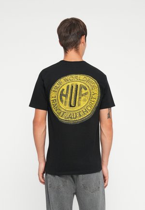 Black cotton t-shirt featuring a large yellow circular logo with "HUF" and text. Short sleeves and crew neckline. Back view shown.