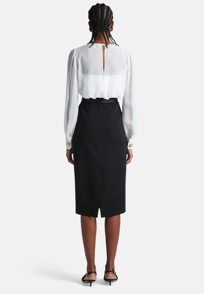 Femme aux cheveux tressés portant une blouse blanche à manches longues et une jupe crayon noire longueur genou, debout de dos sur un fond blanc.