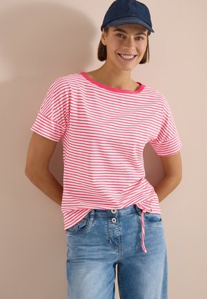 Personne souriante portant une casquette bleue, un t-shirt rayé rose et blanc avec un détail noué sur le côté, et un jean bleu clair, debout contre un mur beige.