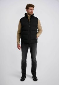 Schwarze Steppweste mit hohem Kragen, Reißverschluss und zwei seitlichen Taschen, getragen über einem hellbeigen Rollkragenpullover, kombiniert mit dunklen Jeans und schwarzen Schuhen.