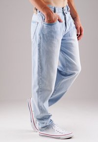 Hellblaue Denim-Jeans mit einem lockeren Schnitt, fünf Taschen und Kupfernieten-Akzenten. Kombiniert mit weißen Sneakern mit roten Streifen.