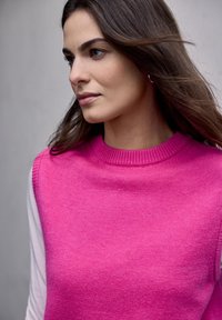 Femme aux longs cheveux bruns portant un pull sans manches rose vif sur un haut blanc à manches longues, regardant sur le côté devant un fond uni.