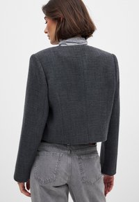 Harmaat lyhyet takki, joka on valmistettu teksturoidusta kankaasta, pitkät hihat ja rakennettu muotoilu. Päällä korkeavyötäröiset harmaat farkut.