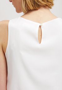 White sleeveless top featuring a keyhole detail at the back, closed with a small button. Smooth fabric with a slight sheen.