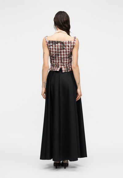 Γυναίκα με σκοτεινά υγρά μαλλιά, που φορά κόσμημα με πέρλες, αμάνικη κόκκινη μπλούζα με καρό μοτίβο, μακριά μαύρη φούστα και ψηλοτάκουνα, στέκεται με την πλάτη.