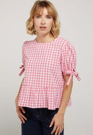 Femme aux cheveux blonds portant un chemisier à carreaux rose et blanc avec des manches bouffantes et des nœuds, associé à un jean bleu foncé.