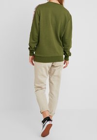 Sudadera verde oliva con dobladillo y puños acanalados; presenta cinta con patrón a lo largo de los hombros. Combinada con pantalones beige ajustados y zapatillas negras.
