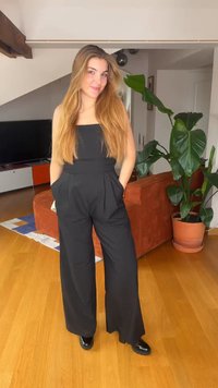 Νεαρή γυναίκα με μακριά ξανθά μαλλιά, φορώντας μαύρη ολόσωμη φόρμα και μαύρα παπούτσια, στέκεται μέσα σε εσωτερικό χώρο κοντά σε πορτοκαλί καναπέ και μεγάλο πράσινο φυτό σε γλάστρα.