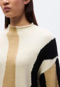 Pull en tricot présentant des motifs abstraits en crème, beige et noir. Texture douce avec un col montant et des épaules tombantes.