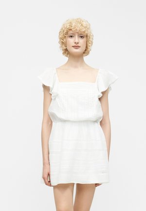 Woman with short curly blonde hair wearing a white short-sleeve dress with flutter sleeves and square neckline, standing against a plain background.