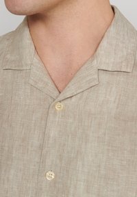 Camicia di lino beige con una texture morbida, colletto corto e patta con due bottoni. Il delicato motivo tessuto migliora il suo aspetto naturale.