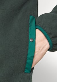 Mørkegrøn fleecejakke med en kontrastfarvet teal lommeforing, der har metaltrykknaplukning og struktureret stof.