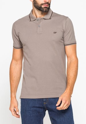 Uomo che indossa una polo a maniche corte marrone chiaro con due bottoni e jeans blu scuro, in piedi davanti a uno sfondo uniforme.