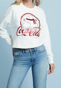 Sweatshirt blanc court avec un logo et une illustration Coca-Cola rouge et noir sur le devant, associé à un jean en denim bleu.