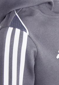 Un sweat à capuche gris avec un motif à trois rayures blanches sur les épaules, présentant un détail en tissu texturé sur le haut des bras et un matériau lisse dans l'ensemble.