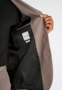 Jasnobrązowa marynarka z satynową czarną podszewką, boczną kieszenią i metką. Gładka tekstura, dopasowany krój i szczegóły z pojedynczym guzikiem.