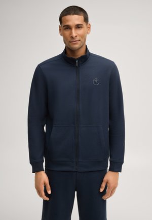 Mann trägt eine marineblaue Jacke mit Reißverschluss, hohem Kragen und vorderen Taschen, steht vor einfachem hellem Hintergrund.