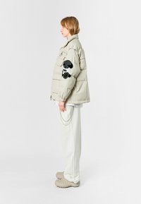 Chaqueta acolchada beige con cierre de cremallera, que presenta un aplique de panda en la manga. Combinada con pantalones blancos y zapatos beige texturizados.