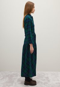 Vestido de cuadros verde y azul marino con mangas largas, cierre de botones en el frente, cintura ceñida y falda amplia. Combinado con zapatos marrones de cordones.