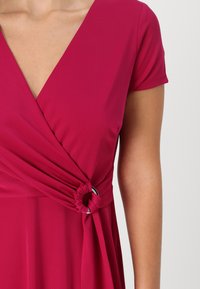 Fuksia-mekko, jossa on V-pääntie ja lyhyet hihat. Ominaisuuksina on kerätty vyötärö solmukydellä ja hopeanvärinen korostus. Silkkinen kangas.