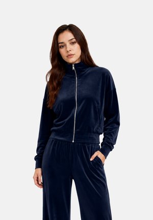 Jeune femme portant une veste zippée en velours bleu marine et un pantalon assorti, une main dans la poche, sur un fond uni.