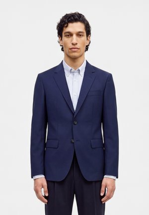 Un homme se tient debout, face à l’avant, vêtu d’un costume bleu marine et d’une chemise bleu clair, sur un fond blanc uni.