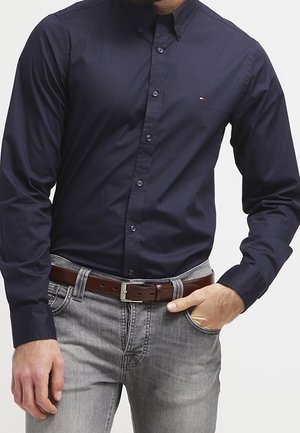 Man draagt een nauwsluitend navy blauw overhemd met knopen, ingestopt in lichtgrijze jeans met een bruine leren riem, hand rust in de zak.