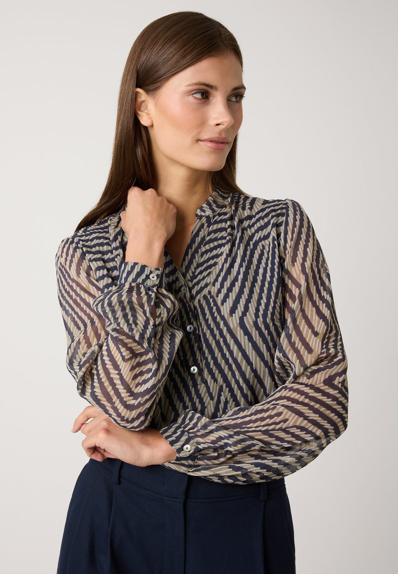 Blouse ample à manches longues en crème avec un motif en zigzag bleu marine, dotée d'une fermeture à boutons à l'avant et de poignets froncés, associée à un pantalon sombre ajusté.