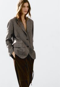 Donna che indossa un blazer grigio con bottoni neri e pantaloni larghi marrone scuro, in piedi con le mani nelle tasche su uno sfondo luminoso e semplice.