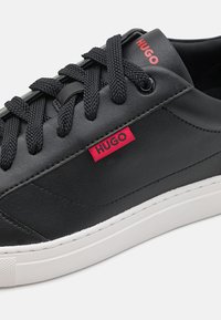 Baskets en cuir noir avec de grosses lacets noirs, semelle blanche, et logos rouges "HUGO" sur la languette et l'étiquette latérale.