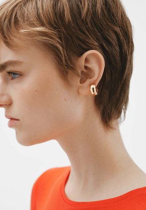 Aro de oro con un diseño moderno, que presenta una superficie lisa y bordes redondeados, usado por un modelo de cabello corto frente a un fondo liso.