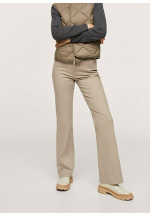 Standing person wearing beige flared pants, cream chunky boots, a quilted brown vest over a dark grey long-sleeve shirt with arms crossed.