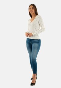 Pulover alb cu guler în V, având un model texturat, cu accente de culoare albastru deschis și margini aurii, asortat cu blugi slim de culoare albastru și pantofi cu toc negri.