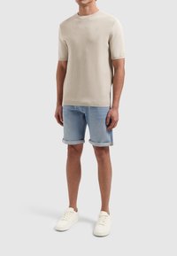 Beige gebreide shirt met korte mouwen en een geribbelde textuur, gecombineerd met lichtblauwe opgerolde denim shorts en witte sneakers.