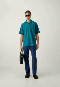 Man som bär en turkos kortärmad skjorta med knappar, mörkblå jeans, svarta skor, solglasögon och håller en svart portfölj mot en neutral bakgrund.