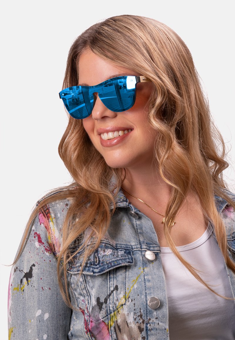 Blue mirrored sunglasses with a geometric frame, gold accents, and a glossy finish, worn with a splatter-painted denim jacket.