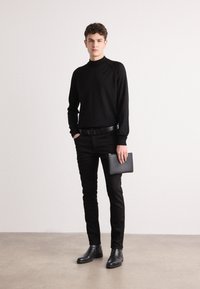 Haut noir à manches longues, pantalon noir ajusté, ceinture noire et bottines. Tenant une pochette noire, sur un fond neutre.