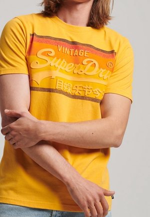 Personne portant un t-shirt vintage SuperDry jaune avec des rayures orange et marron, croisant le bras gauche sur le bras droit.