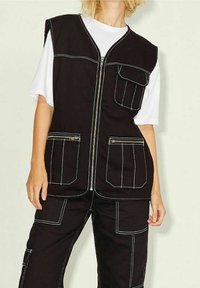 Svart väst med vit söm, med en framdragkedja, två stora framfickor och en avslappnad passform. Buren över en vit skjorta.