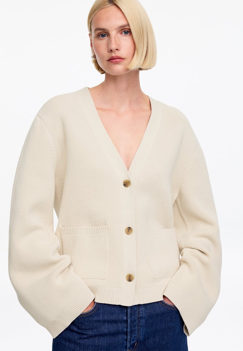 Jeune femme aux cheveux blonds courts porte un cardigan crème oversize avec trois boutons, poches avant, et jean bleu, mains dans les poches.