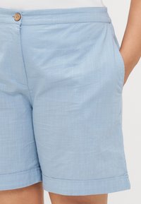 Pantalones cortos de lino de color azul claro con una textura suave, que presentan bolsillos laterales, un cierre de botón y dobladillos enrollados para un diseño informal.
