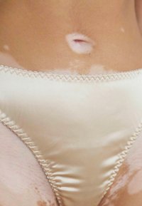 Bas de bikini en satin beige avec un bord festonné, mettant en valeur la peau avec des tons clairs et foncés. Présente un détail circulaire au centre.