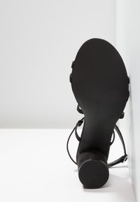 Chaussure à talon haut noire avec une semelle texturée, en matériau suédé, et de fines brides pour la cheville. Le talon a une forme cylindrique avec une base solide.