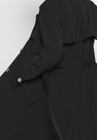 Manteau trench noir à silhouette évasée, détail de boutons à l'avant, et ceinture. Présente un col à couches et une texture lisse.